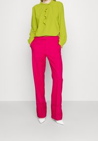Grøn bluse med flæse-detaljer, parret med lyserøde bukser. Hvide spidse hæle komplementerer det livlige outfit. Glat stoffinish.