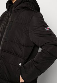 Giacca a vento nera con cappuccio; presenta un design trapuntato, chiusura con zip frontale, tasche laterali e una toppa con logo sulla manica. Materiale morbido e testurizzato.