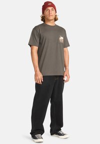 Camiseta de algodón gris con mangas cortas y un bolsillo en el pecho que presenta un diseño estampado. Combinada con pantalones negros de corte holgado y zapatillas de deporte azul marino.