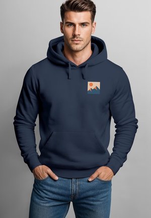 Junger Mann trägt einen dunkelblauen Hoodie mit einer kleinen Berg- und Sonnengrafik auf der Brust, steht mit den Händen in den Taschen.