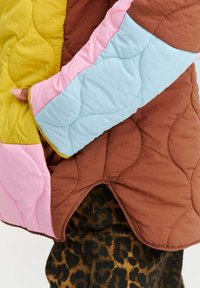 Osoba oblečená v prešívanej bunde s hnedými, ružovými, modrými a žltými panelmi, drží okraj bundy a pod ňou sú viditeľné nohavice s leopardím vzorom.