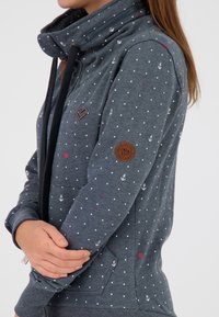 Felpa grigia con pois bianchi e rosa, caratterizzata da motivi di ancore, colletto alto e dettagli con zip. Include un emblema a forma di cuore e una patch in pelle.
