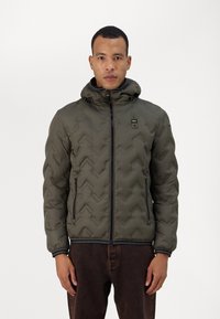 Giacca trapuntata verde con un motivo a chevron, apertura con zip e cappuccio. Presenta cerniere nere e una toppa con logo sul petto. Texture liscia.