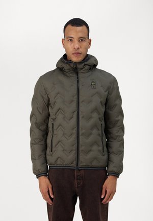 Chaqueta acolchada verde con un patrón en forma de chevron, con cierre frontal y capucha. Presenta cremalleras negras y un parche de logo en el pecho. Textura suave.