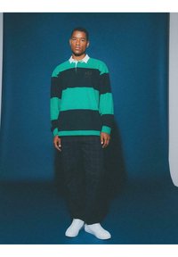 Zielono-czarny rugby z paskami, z białym kołnierzykiem, sparowany z ciemnymi spodniami w kratę i białymi trampkami. Gładkie tło z cieniami.