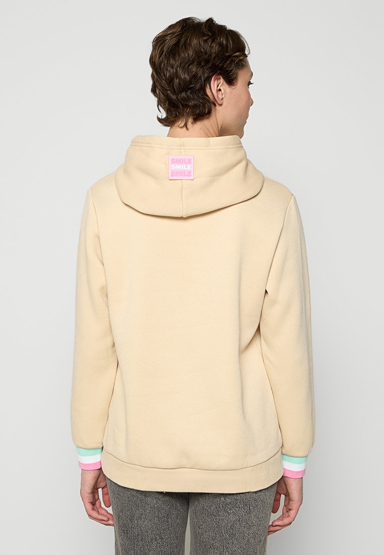 Beżowa bluza z kapturem o miękkiej teksturze. Posiada kolorowe ściągacze w różowym, miętowym i jasnoniebieskim kolorze. Różowa metka z tyłu.