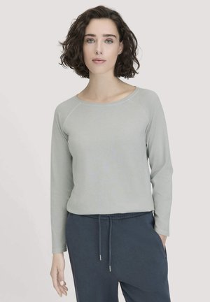Frau mit kurzen lockigen dunklen Haaren, die ein hellgraues Langarmshirt und dunkelblaue Kordelhose trägt, steht vor einem neutralen Hintergrund.