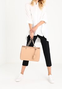 Sac fourre-tout beige avec anses en chaîne argentée, forme rectangulaire, texture en cuir lisse, petit détail d'étiquette, associé à un pantalon noir et des baskets blanches.