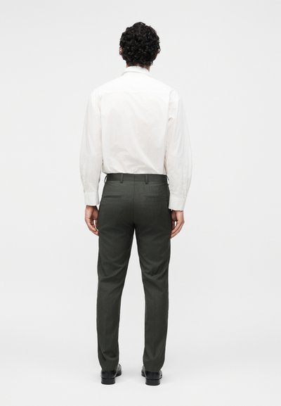 Άνδρας με σγουρά μαλλιά που φοράει λευκό πουκάμισο με μακριά μανίκια, σκούρο γκρι παντελόνι και μαύρα παπούτσια, στέκεται με την πλάτη προς ένα απλό φόντο.