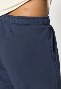 Tumši zilie treniņbikses ar gludu tekstūru, elastīgu jostu un sānu kabatām. Iespējama neliela spīdēšana un bez redzamām rakstiem.