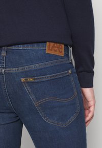 Jean en denim bleu foncé avec une finition texturée. Étiquette en cuir marron à la taille avec le logo « Lee ». Couture contrastée sur les poches.
