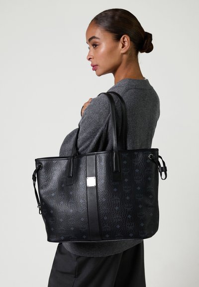 Donna in maglione grigio tiene una grande borsa tote nera con texture e dettagli a motivo e doppie spalline.