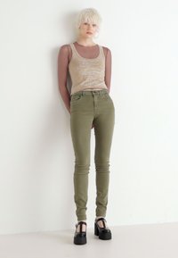 Top sin mangas beige claro, jeans ajustados verde oliva y zapatos de plataforma negros. Diseño sencillo, forma ajustada, textura suave y detalles sutiles.