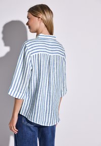 Camicia a maniche corte a righe blu e bianche con una piega sul retro, realizzata in tessuto leggero. Presenta un colletto standard e fessure laterali.