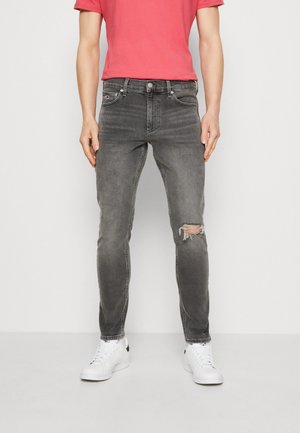 Mann trägt ausgewaschene schwarze zerrissene Skinny-Jeans mit weißen Sneakern und einem korallenfarbenen kurzärmligen Hemd, steht vor einem schlichten weißen Hintergrund.