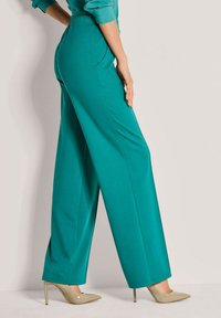 Vrouw in teal kleurige wijde broek en beige hoge hakken, staand tegen een lichte achtergrond met één hand op haar dij rustend.