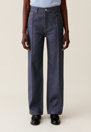 Osoba nosząca ciemnoniebieskie, proste jeansy z wysokim stanem i przednimi szwami, w połączeniu z czarnymi loafersami i jasnoniebieskim topem.