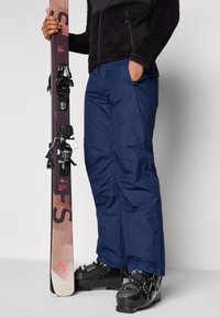Skiausrüstung bestehend aus marineblauen wasserdichten Hosen, einer schwarzen Fleecejacke, schwarzen Skischuhen mit grünen Akzenten und Skiern mit Holz- und lila Design.