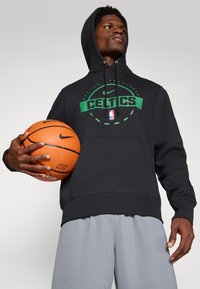 Čierna mikina s kapucňou a zeleným logom "Celtics", obsahujúca klokaniu kapsu a šnúrku na kapucni. Sivé šortky; drží oranžovú basketbalovú loptu Nike.
