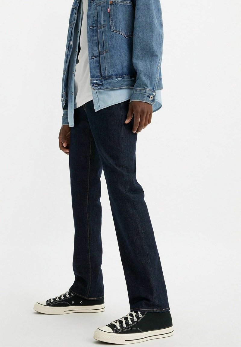 Homme portant une veste en jean superposée, une chemise blanche, un jean foncé et des baskets noires et blanches, debout devant un fond uni.
