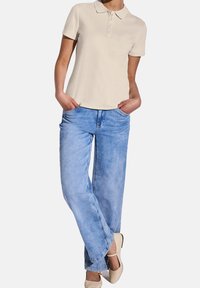 Polo beige avec un col et cinq boutons, associé à un jean en denim large de couleur bleu clair. Le modèle se tient avec les mains dans les poches.