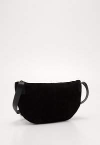 Černá semišová taška s zakřiveným tvarem, zipovým uzávěrem a plochým koženým popruhem. Má hladkou texturu a minimalistický design.