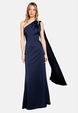 Donna che indossa un abito da sera in raso blu navy monospalla con un lungo tessuto drappeggiato che si estende dalla spalla, in piedi davanti a uno sfondo semplice.