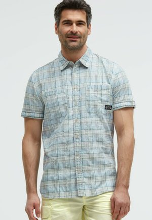 Mann trägt ein kurzärmeliges, hellblau kariertes Hemd mit Knöpfen und hellgelbe Shorts vor einfachem Hintergrund.