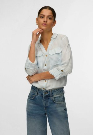 Kvinde poserende med hånd på hagen, iført op-rullede, stribede skjorte og blå denimjeans mod en ensfarvet baggrund.