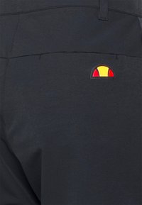 Pantalon bleu foncé avec une poche arrière portant un petit logo circulaire rouge et jaune brodé près de la taille. Texture lisse.