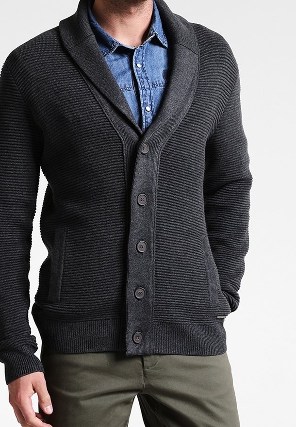 Svart ribbad cardigan med sjalkrage, stora knappar och två framfickor, buren över en denimskjorta. Texturerat stickat tyg.