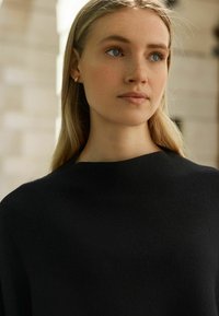 Svart stickad tröja med hög, rundad halsringning. Slät textur och minimala design detaljer, med en närbild av halsringningen och kragen.