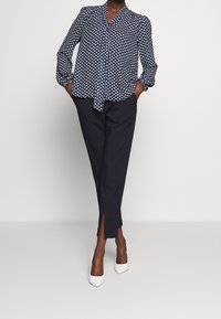 Blusa navy con motivo a cuori bianchi, con nodo frontale e maniche a sbuffo lunghe; abbinata a pantaloni neri sartoriali con aperture laterali. Tacchi bianchi alti.