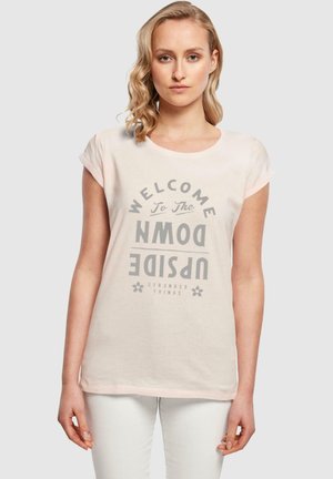 Lys rosa bomulls T-skjorte med rullet korte ermer. Har grå grafisk tekst "VELKOMMEN TIL DEN OPPNED VERSEN" fra "Stranger Things."