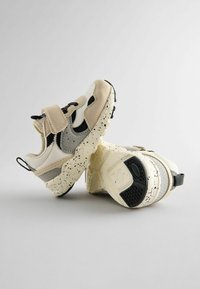 Baskets blanches et beiges avec accents gris, semelle mouchetée, bande velcro et matériaux texturés. Comprend une languette au talon.