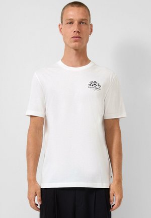 Mann trägt ein weißes T-Shirt mit kurzen Ärmeln, schwarzem Text und Grafik auf der linken Brust, kombiniert mit einer schwarzen Hose.