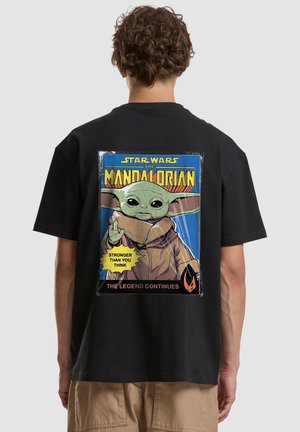 Person trägt schwarzes T-Shirt mit Star Wars The Mandalorian Grafik, die Baby Yoda und den Text "Stronger than you think." zeigt