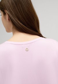 Lichtroze top met een ronde halslijn, zachte textuur en een klein logo-accent op de achterkant. Eenvoudig en minimalistisch ontwerp.