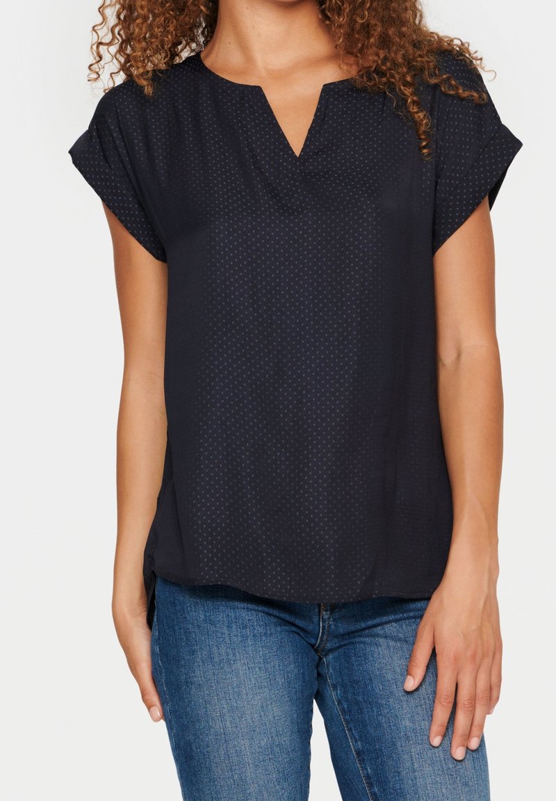 Haut bleu marine à manches courtes en tissu léger avec un col en V et un motif à pois subtil, associé à un jean en denim. Texture douce et coupe décontractée.