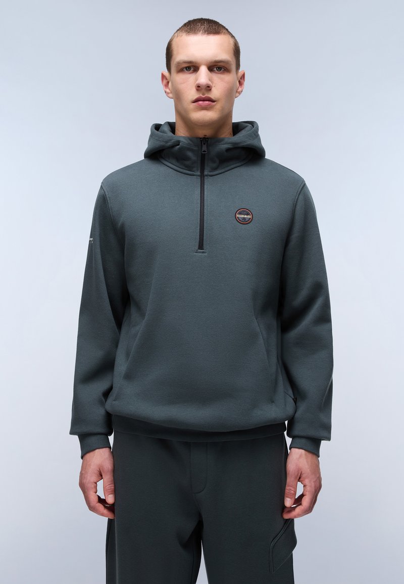 Mørkegrøn hoodie lavet af blødt stof, med en halvlukket design, forreste logo-patch og justerbare snore i hætten.