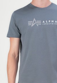 Pilkas medvilninis marškinėliai su apvaliu kaklu, trumpomis rankovėmis ir ryškiu baltu bei šviesiai pilku logo, kuriame matomi abstraktūs linijų ir teksto motyvai.