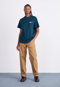 Dark teal cotton t-shirt with a crew neckline and short sleeves, paired with light brown trousers and black platform shoes. Minimalist design.