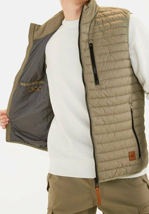 Homme portant un gilet matelassé beige sur une chemise blanche à manches longues, tenant le gilet ouvert pour montrer la doublure intérieure foncée et les détails des poches.