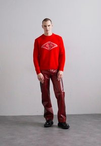 Röd stickad tröja med vit diamantgrafik, matchad med glänsande burgundybyxor och svarta chunky-skor, stående mot en neutral bakgrund.