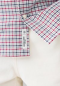 Karirani košulja u crvenoj, mornarskoj i bijeloj boji od pamuka s kragnom na gumb i oznakom "Durable Goods Lot 082." Pozadina je bijela.