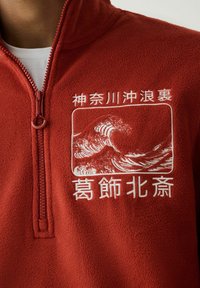 Közeli kép egy piros polárról, amelyen negyedcipzár és a mellrészen fehér japán felirat valamint hullám mintázat hímzés látható.