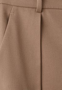 Pantalons beiges en tissu texturé, avec un devant plat comportant des plis cousus, et une taille épurée sans fermetures visibles.