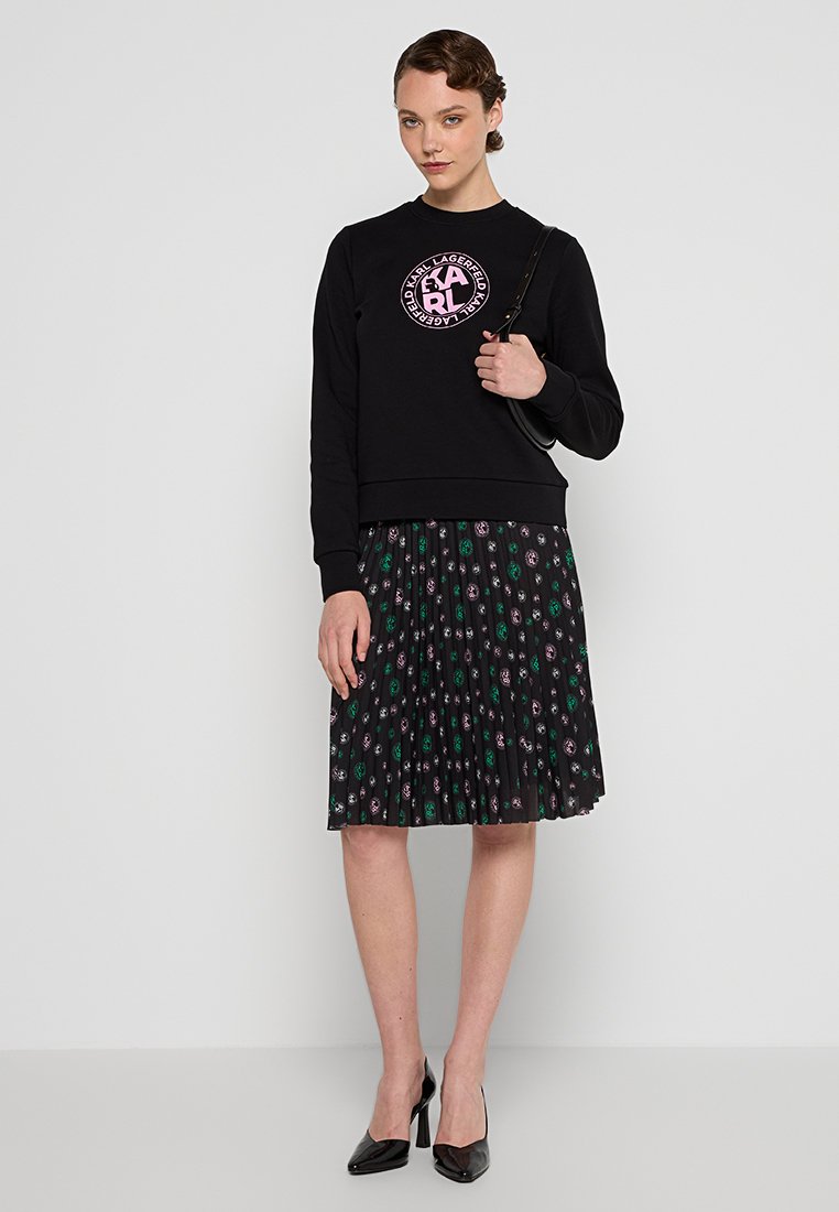 Mujer de pie vestida con un sudadera negra con un logo rosa, falda plisada negra con patrones morados y verdes, tacones negros y sosteniendo un bolso negro.