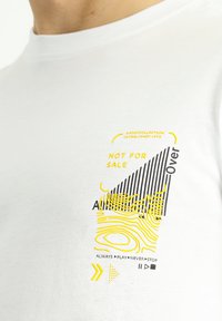 Camiseta blanca de algodón con un diseño gráfico geométrico en negro y amarillo, que incluye elementos de texto y patrones de ondas en el frente.
