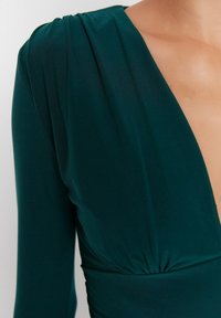 Robe bleu profonds avec décolleté plongeant, manches longues et détails froncés à la taille. Tissu lisse et extensible avec un léger relief.